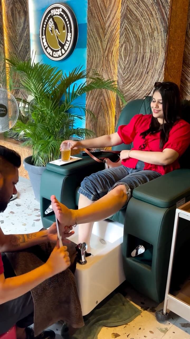 A guest in a pedicure chair with a cold drink, a therapist working at her feet, the studio’s badge on the wall behind